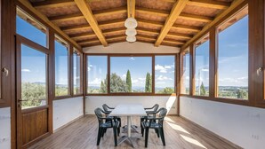 Amplia habitación con una mesa grande y sillas, vigas de madera en el techo y ventanas amplias que ofrecen vistas a árboles y colinas.
