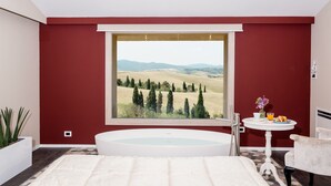 A bedroom with a large window, a bathtub, a small table with a vase and a glass of orange juice, and a chair.