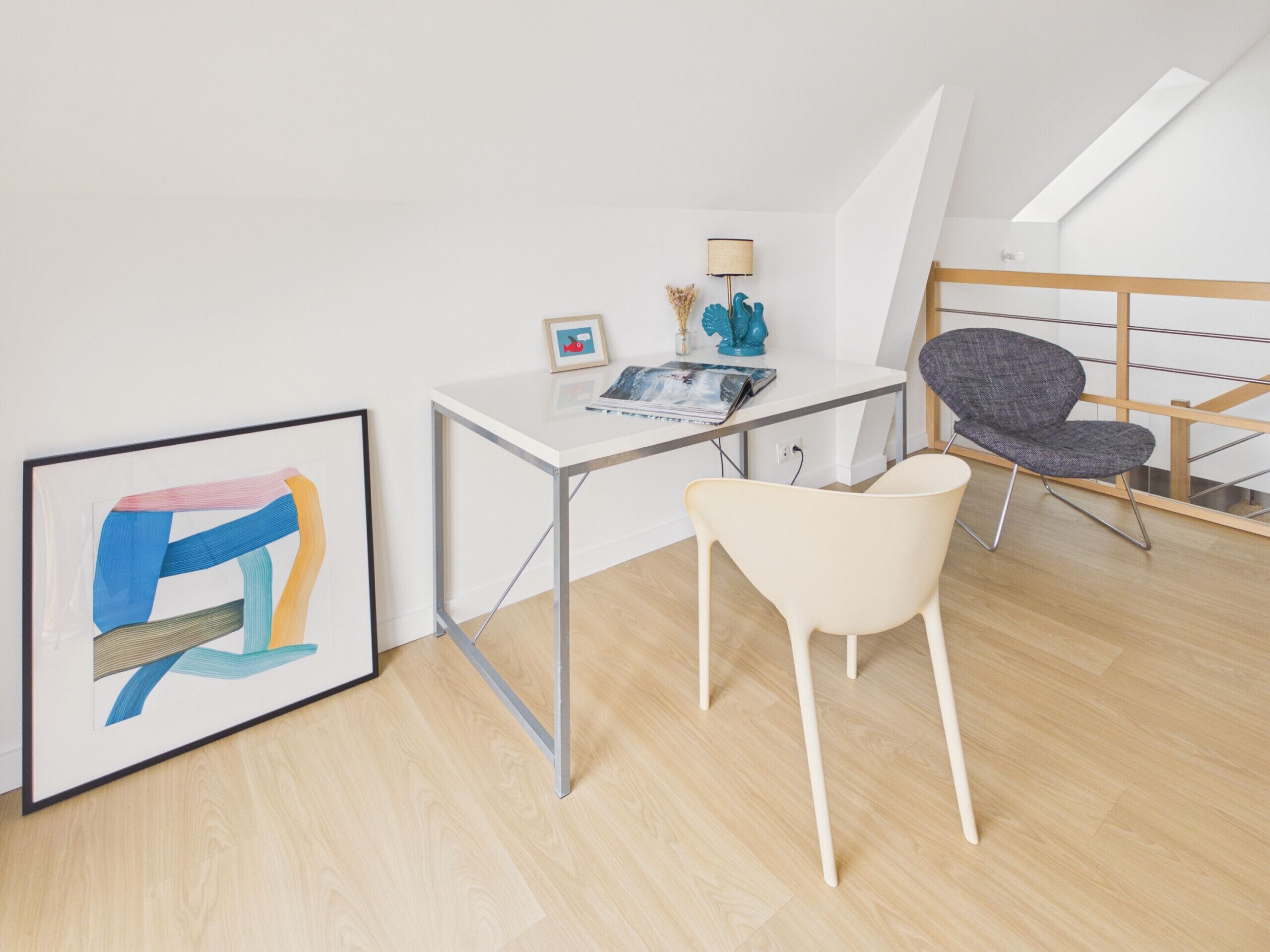 A modern room with a desk, a chair, a framed abstract painting, a lamp, and a staircase.
