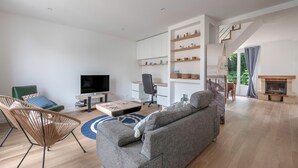 Un salon moderne comprenant un canapé, un fauteuil, un téléviseur, un bureau et des étagères ornées d’objets décoratifs.
