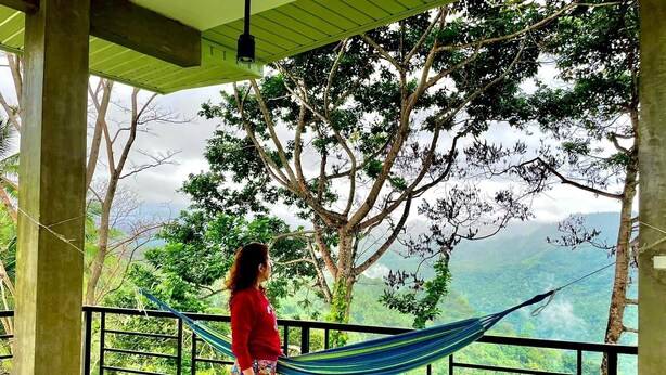 A person standing on a balcony with a hammock, overlooking a lush green landscape.