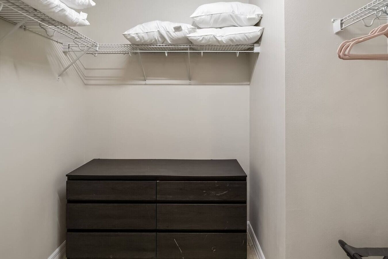 Un petit meuble de rangement de couleur sombre, doté de plusieurs tiroirs, une étagère en métal au-dessus où sont posés des coussins blancs, et un étendoir métallique avec des cintres sur le côté droit.