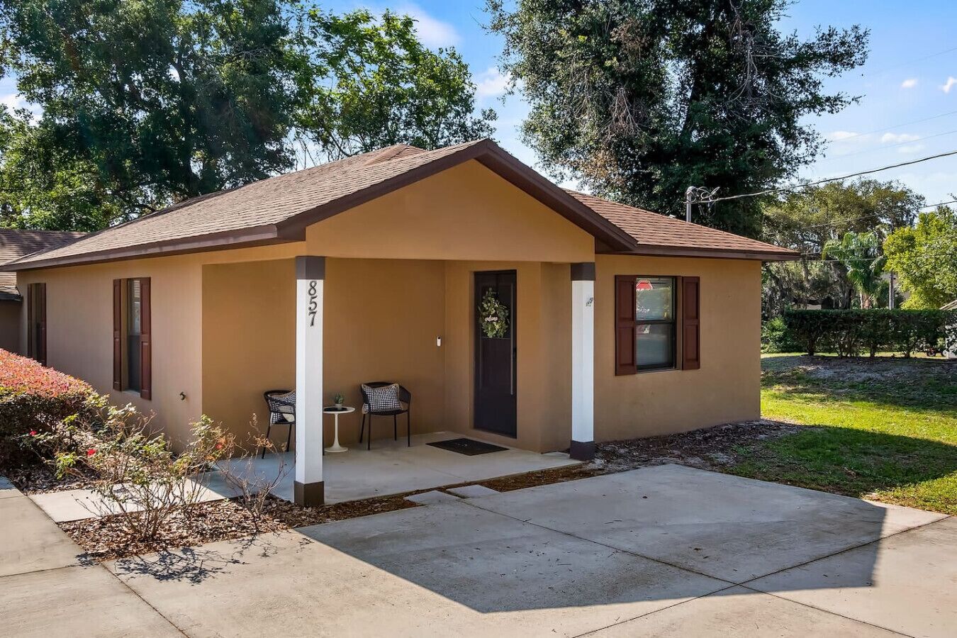 A small, single-story building with a brown roof and beige walls, featuring a porch with two chairs and a small table.