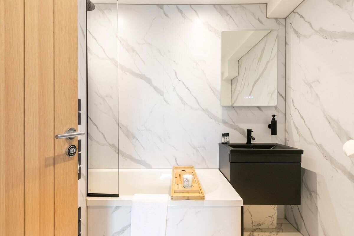 A modern bathroom with a marble wall, a wooden door, a black sink, and a wooden tray on the counter.