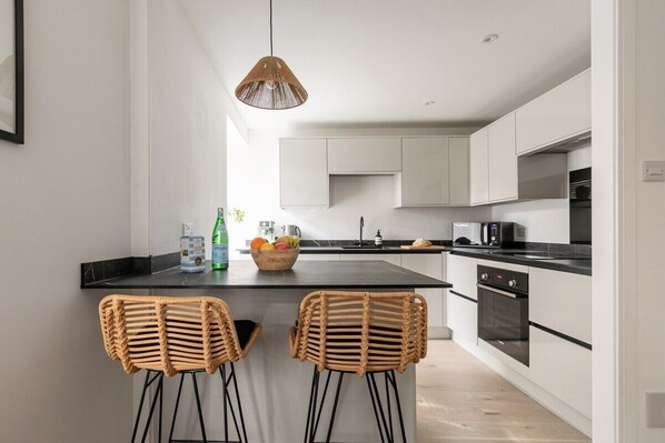A modern kitchen with a central island, two wicker chairs, a hanging light fixture, and built-in appliances.