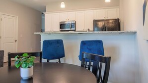A modern kitchen with white cabinets, a stainless steel refrigerator, and a microwave. There are two blue chairs around a round table with a potted plant. - Feels Like Home! Columbia near Fort Jackson - Perfect for Families - Sleeps 6 (Elgin)