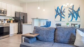 A modern kitchen with white cabinets, stainless steel appliances, and a dining area with a gray sofa and blue artwork. - Feels Like Home! Columbia near Fort Jackson - Perfect for Families - Sleeps 6 (Elgin)