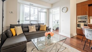 A modern living room with a glass coffee table, a dark sofa with patterned cushions, and a view of the city through large windows.
