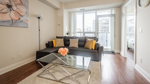 A modern living room with a glass coffee table, a sofa with cushions, a large window, and a painting on the wall.