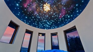 Una habitación con techo estrellado, tres ventanas con vistas panorámicas y una cama con manta azul.