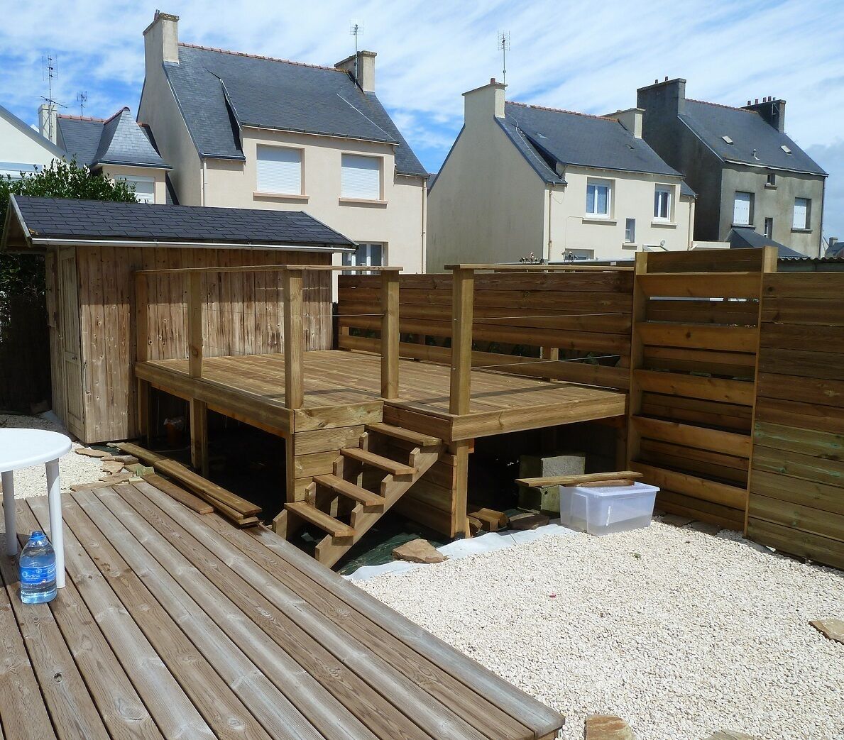 Une terrasse en bois avec un escalier menant à une plateforme surélevée, le tout entouré d’une clôture en bois, avec des maisons en arrière-plan.