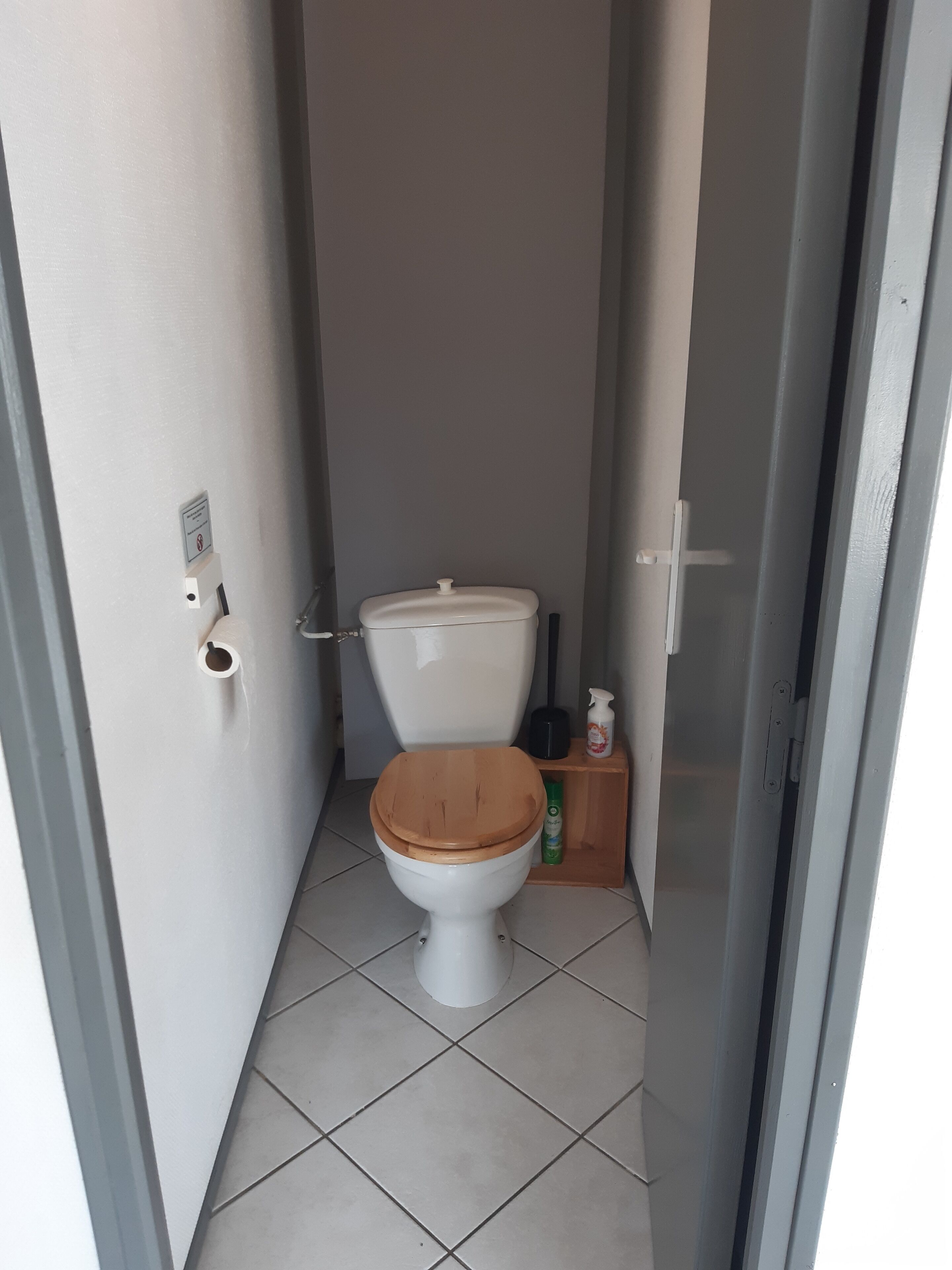 A small, narrow bathroom with a white toilet, a wooden seat, and a small shelf with toiletries.