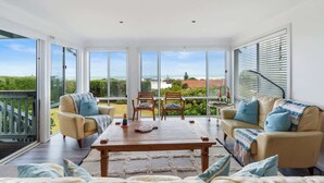 Een ruime woonkamer met een grote schuifpui naar een balkon, een houten salontafel, twee beige banken met blauwe kussens, en uitzicht op de oceaan en groen.