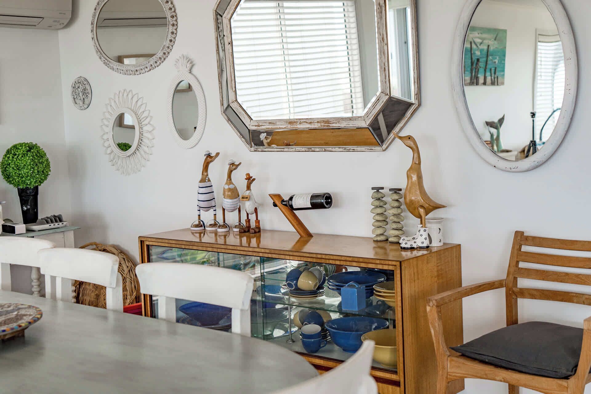 Un coin repas comprenant une table en bois, des chaises, un meuble vitré où sont exposées de la vaisselle, et plusieurs miroirs muraux.