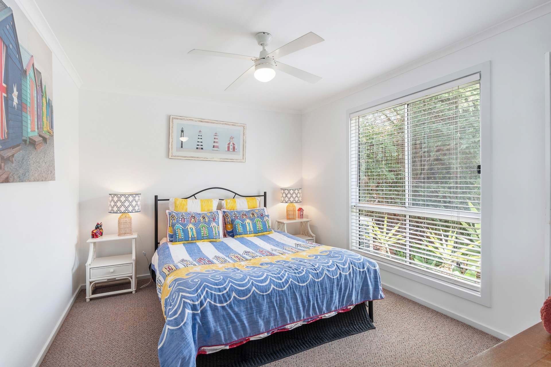 Une chambre à coucher comprenant un lit, deux tables de chevet, un ventilateur de plafond, une fenêtre avec des stores et un tableau accroché au mur.
