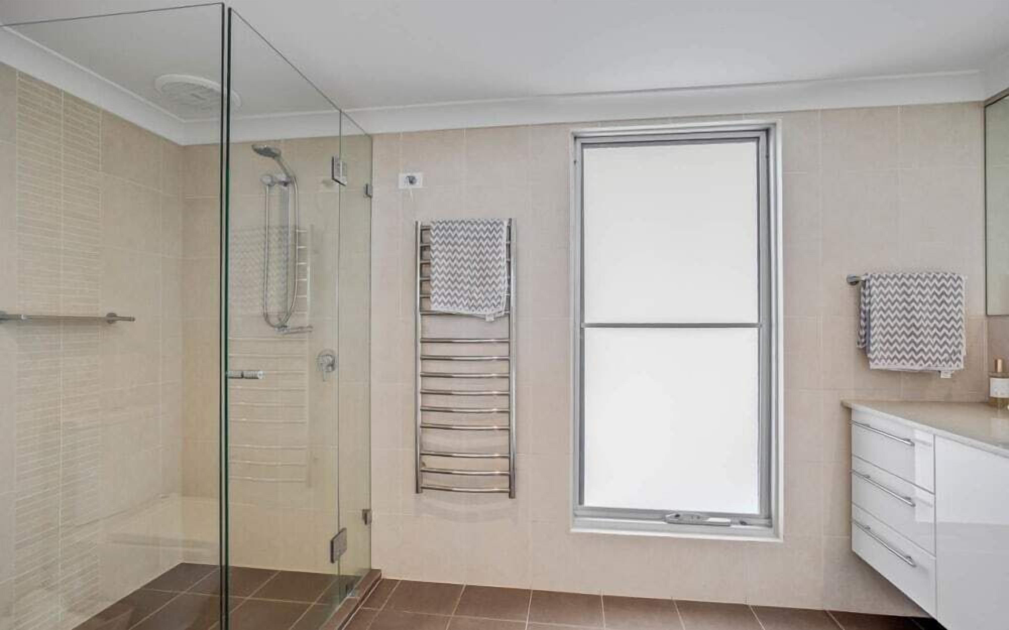 Un baño moderno con cabina de ducha de vidrio, un ventanal grande y un mueble de tocador blanco con cajones.