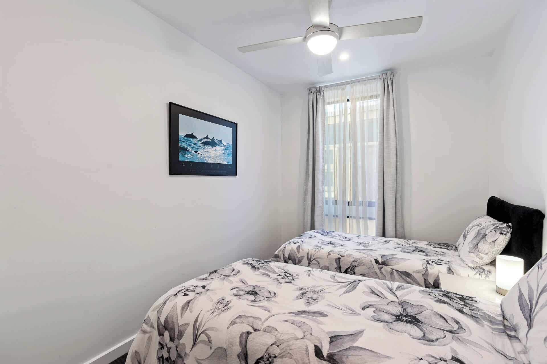 Un dormitorio con dos camas, un ventilador de techo, una ventana con cortinas e imagen enmarcadas en la pared.