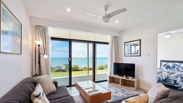 A modern living room with a sofa, TV, and a view of the sea through large glass doors. - The Dolphins 1 – Beachfront Holiday Apartment | 1/10 Strangways Terrace (Port Elliot)
