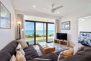 A modern living room with a sofa, TV, and a view of the sea through large glass doors. - The Dolphins 1 – Beachfront Holiday Apartment | 1/10 Strangways Terrace (Port Elliot)