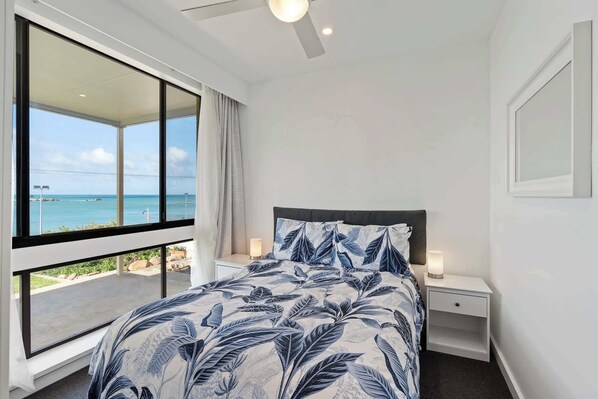 A bedroom with a large window, a bed with a leaf-patterned bedspread, two bedside tables with lamps, and a view of the ocean. - The Dolphins 1 – Beachfront Holiday Apartment | 1/10 Strangways Terrace (Port Elliot)