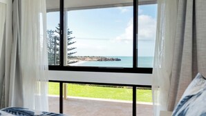 A bedroom with a large window overlooking a rocky coastline and a grassy area. - The Dolphins 1 – Beachfront Holiday Apartment | 1/10 Strangways Terrace (Port Elliot)