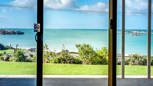 A view through a glass door of a coastal area with a clear sky, a calm sea, and rocky outcrops. - The Dolphins 1 – Beachfront Holiday Apartment | 1/10 Strangways Terrace (Port Elliot)