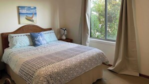 Une chambre à coucher avec un lit, une table de chevet, une lampe, une fenêtre avec des rideaux et un tableau au mur.