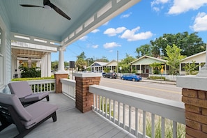 A porch with two lounge chairs, a ceiling fan, and a view of a street with houses and cars.