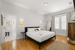 Une chambre avec un plancher en bois, un lit avec une literie blanche, deux lampes de chevet et une commode en bois.