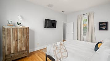 Une chambre avec une armoire en bois, un lit recouvert d’une courtepointe blanche, un téléviseur fixé au mur et une fenêtre avec des rideaux.