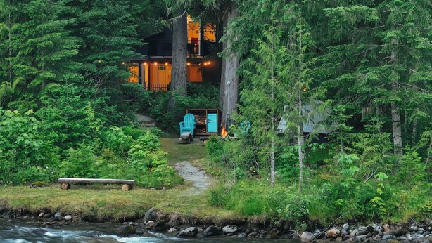 Una cabaña rodeada de un bosque frondoso, con una terraza de madera y un río.