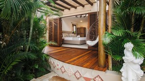Terraza de madera con una cama, rodeada de exuberante vegetación y una estatua.