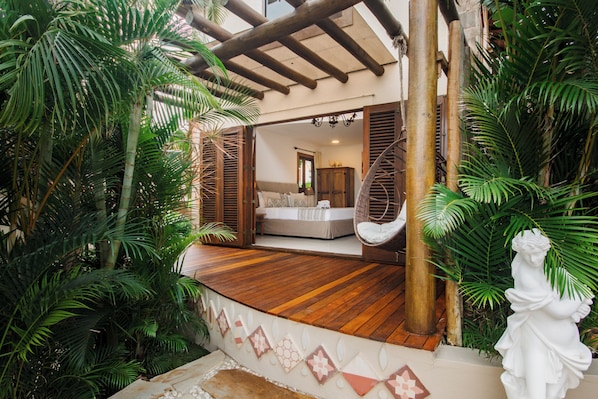 Terraza de madera con una cama, rodeada de exuberante vegetación y una estatua.