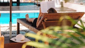 Una persona descansando en una silla de madera junto a una piscina, con un libro, rodeada de sombrillas y una estatua.