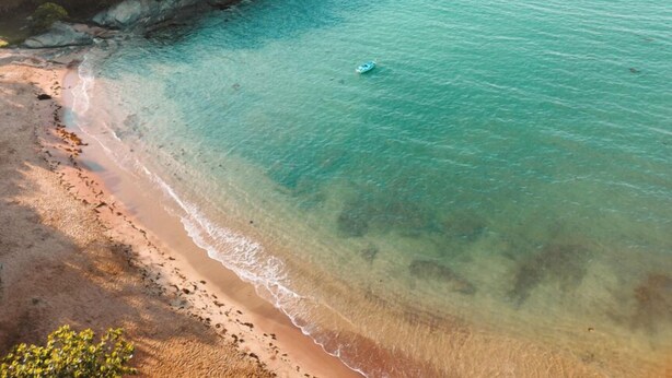 Vista aérea de una zona costera con playa, aguas turquesas cristalinas y una pequeña casa rodeada de vegetación.