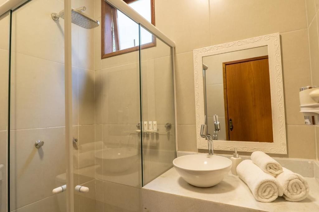 Baño con ducha de mampara de vidrio, lavabo redondo y espejo con reflejo de una puerta de madera.