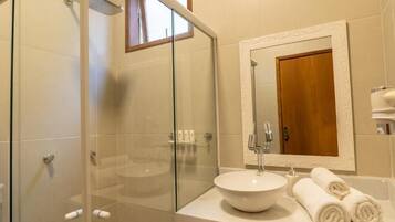 Baño con ducha de mampara de vidrio, lavabo redondo y espejo con reflejo de una puerta de madera.