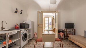 Een kleine kamer met een wasmachine, een wastafel, een klein tafeltje met stoelen, een bed en een televisie.