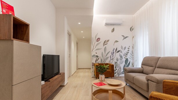 A modern living room with a sofa, a TV on a wooden stand, a small table with a plant, and a wall with a leaf pattern. - Santamaria Apartment by Wonderful Italy (Siracusa)