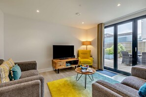 A modern living room with a flat-screen TV, a yellow armchair, and a dining area with wicker furniture. - 2 Bed in Woolacombe (oc-b29816) (Woolacombe)