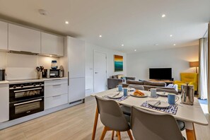 Une cuisine moderne dotée d’appareils électroménagers intégrés, un coin repas avec une table dressée pour deux personnes, et un salon avec un canapé et un téléviseur.