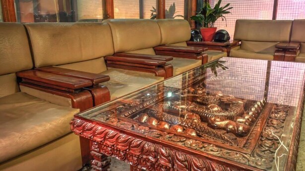 A living room with a glass-top coffee table featuring an intricate metal sculpture, leather sofas, and wooden side tables. - Hotel Kamal (Kathmandu)
