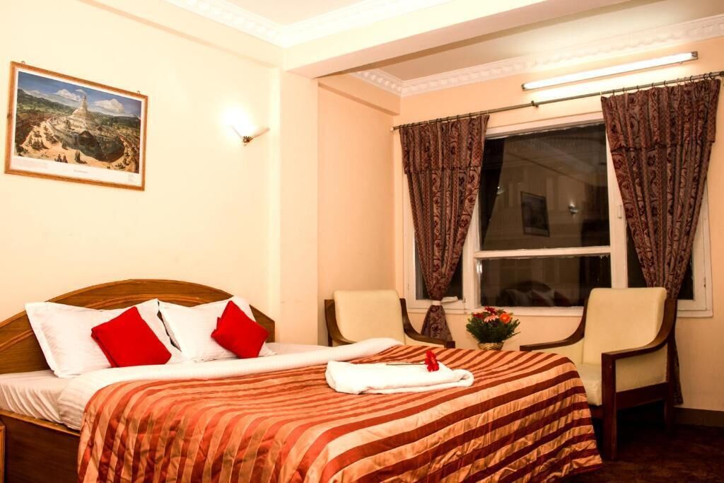 Ein Hotelzimmer mit Bett, zwei Stühlen, einem Fenster mit Vorhängen, einem Bild an der Wand und einem kleinen Tisch mit einer Blume.