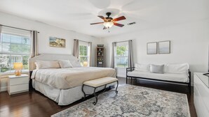 A bedroom with a bed, bedside tables, a bench, a ceiling fan, and a patterned rug. - Oak trees, Heated Pool oasis & walk to beach! Drive to downtown 3 min! (Gulfport)