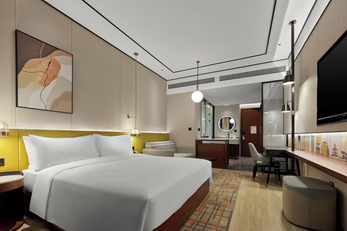 A modern hotel room with a large bed, a desk with a chair, a mirror, and a TV.