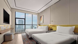 A modern hotel room with two beds, a large window offering a city view, a flat-screen TV, and a small trash bin.