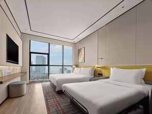 A modern hotel room with two beds, a large window offering a city view, a flat-screen TV, and a small trash bin.