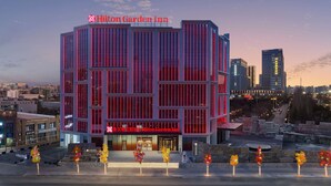 A modern Hilton Garden Inn with a prominent red sign, large windows, and a view of the cityscape in the background.