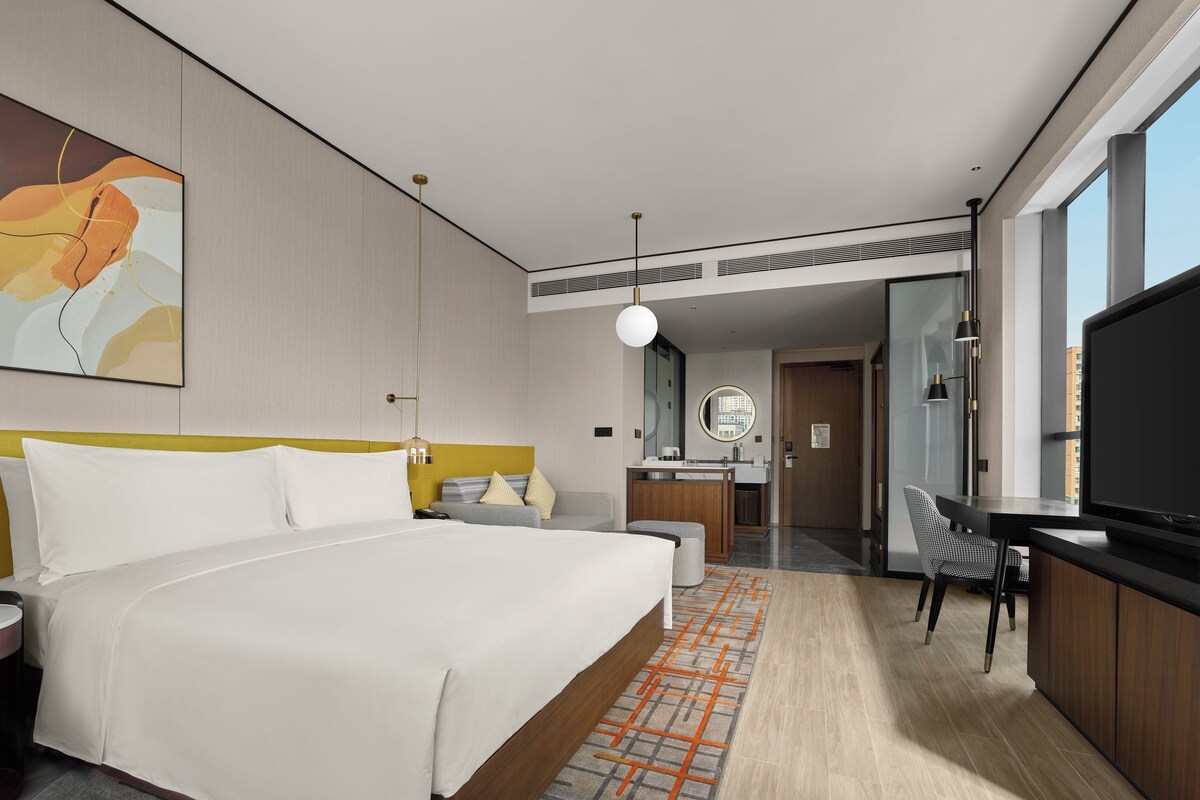 A modern hotel room with a large bed, a desk, a chair, a TV, and a bathroom visible through an open door.