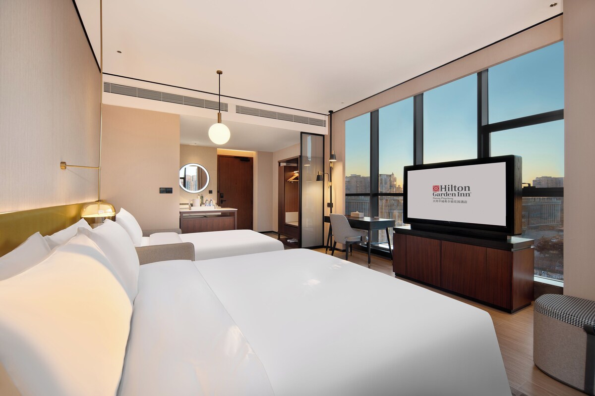 A modern hotel room with a large bed, a desk, a chair, a TV, and a view of the city.